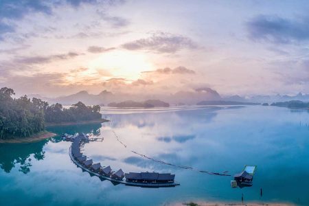 serene view of Khao Sok Lake at dawn, with mist floating above the calm waters, creating a mystical and ethereal atmosphere.